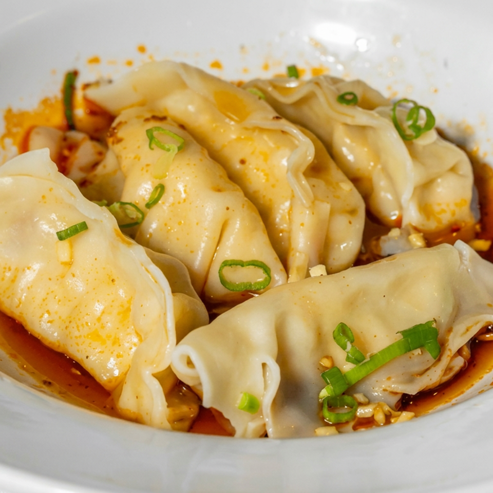 Steamed dumplings topped with green onions, served in spicy red chili oil on a white plate.