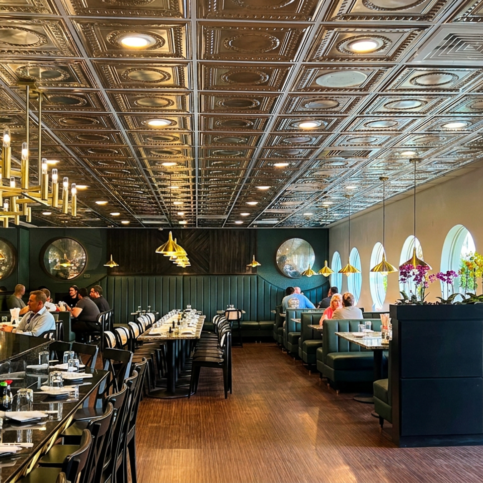Elegant restaurant dining room with ornate tin ceiling, brass chandeliers, dark green booths, marble bar, plants, and seated diners.